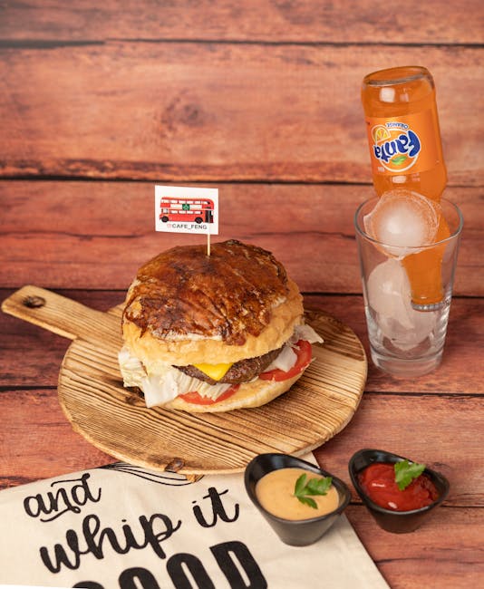 A classic hamburger steak sandwich with a toasted bun, filled with fresh lettuce, tomato slices, a beef patty, melted cheese, and a fried egg on top. It rests on a round wooden serving board. Next to