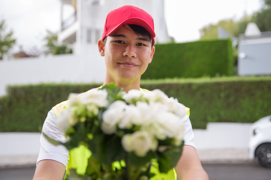 A delivery person wearing a red cap and a neon yellow vest is smiling and holding a bouquet of fresh white flowers with green leaves, extending it towards the camera in an outdoor setting. The bouquet