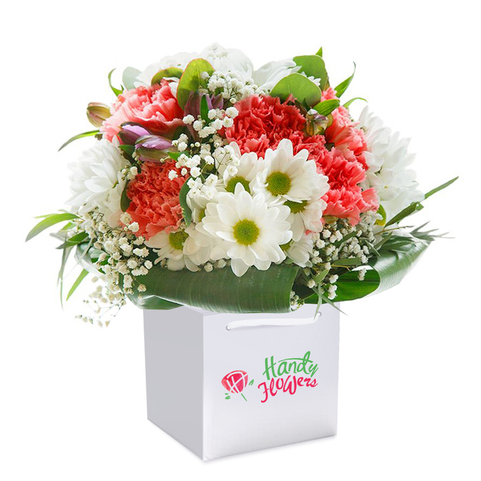 Set against the purest white background, this striking floral bouquet is arranged upright in a modern, crisp white square gift bag-an emblem of Penge's blend of contemporary flair and community warmth. The main event is a harmony of lush, coral-hued carnations with their signature soft, ruffled petals, paired elegantly with snowy white daisies crowned by zingy lime-green centers. Wisps of white baby's breath give the ensemble an ethereal quality, as if caught in a gentle breeze swirling through nearby Cator Park. The inclusion of unopened blooms tinted with purple hints suggests growth and hope, artfully breaking up the rounded silhouette and inviting closer admiration. Encircling the flowers, a collar of deep, glistening foliage echoes the verdant lawns of Penge's local gardens, providing essential structure and a sense of natural abundance. The sturdy ribbon handles and clean, minimalist design of the gift bag demonstrate both practicality and attention to detail-hallmarks of a local florist's care. This composition radiates freshness and thoughtful beauty, perfect for occasions from birthdays to 'thinking of you' deliveries across SE20. Its ready-to-gift presentation ensures every recipient experiences a sense of delight and anticipation upon its arrival at their Penge home.
