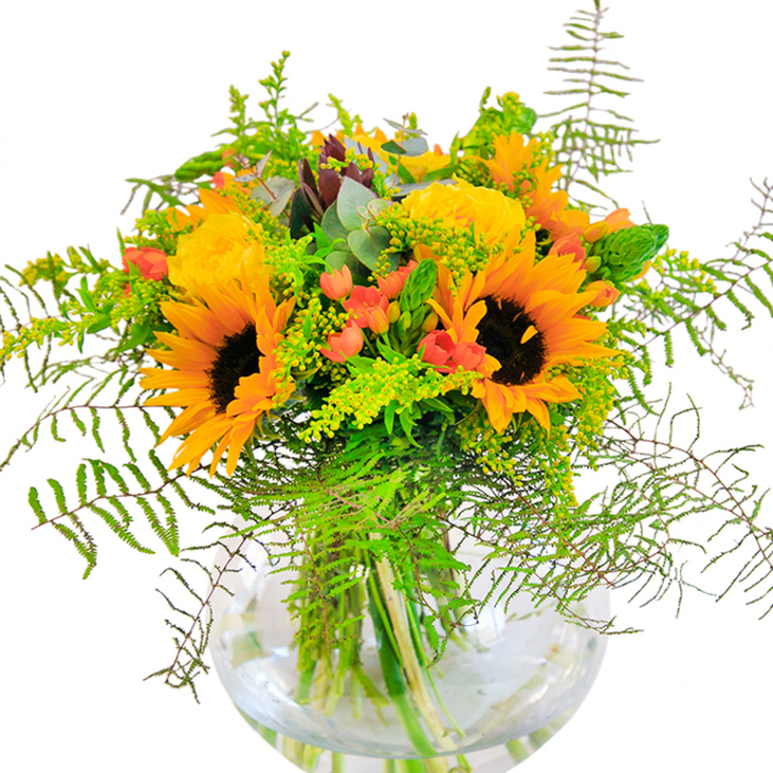 This hand-curated bouquet, inspired by the vibrant character of Penge, radiates pure joy and warmth from the moment you encounter it. Sunflowers take centre stage, stretching out golden petals like rays over Melvin Road in full summer bloom. Their deep brown centres, rich and velvety, ground the arrangement and serve as a reminder of nature's abundance. Around them, buttery yellow garden roses open in layers, each petal tightly packed in an elegant whirl-echoing the classical gardens tucked behind Penge's Victorian terraces. Delicate clusters of ruby-red Hypericum berries offer sparkling contrast, nestled amid generous greenery. Feathery fronds of fern spill gracefully, imitating the movement of leaves stirred by a gentle breeze in Crystal Palace Park, while solidago's tiny yellow flowers add bursts of brightness between the larger blooms. Silvery blue eucalyptus leaves peep through, softly perfumed and cool in hue. Held within a clear, sphere-like vase, all the vibrant stems are visible, their fresh cut ends awash in clean water, enhancing the clean, contemporary feel. The overall effect is both striking and harmonious: a celebration of Penge's welcoming, close-knit spirit, perfectly crafted by Florist Penge for special occasions-a thoughtful gesture for anniversaries, birthdays, or simply to bring a piece of sunlight indoors. The bouquet's textures invite touch and its colours evoke the happiest days of the year.